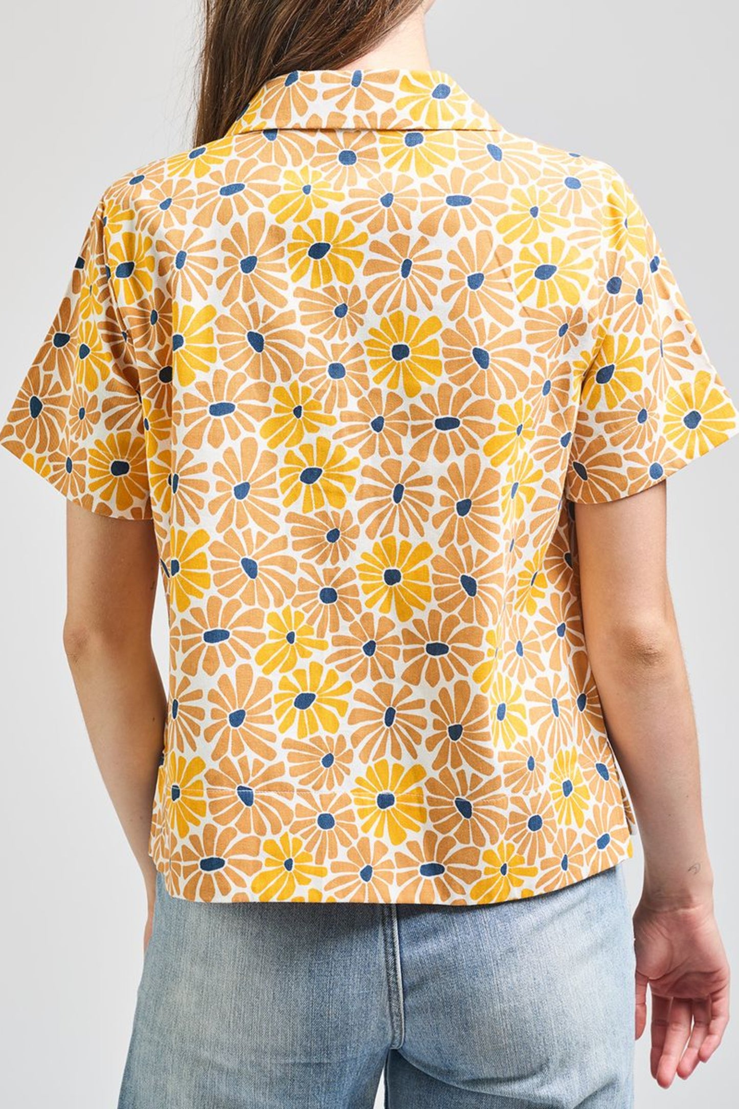 Image of model wearing MATA TRADERS Fair Trade Certified Camp Shirt in Sunny Floral Print. Camp Top is an open neck vintage inspired short sleeve button down. Sunny Floral Print is an orange, yellow, and teal sunflower motif hand block printed on Cotton twill fabric.