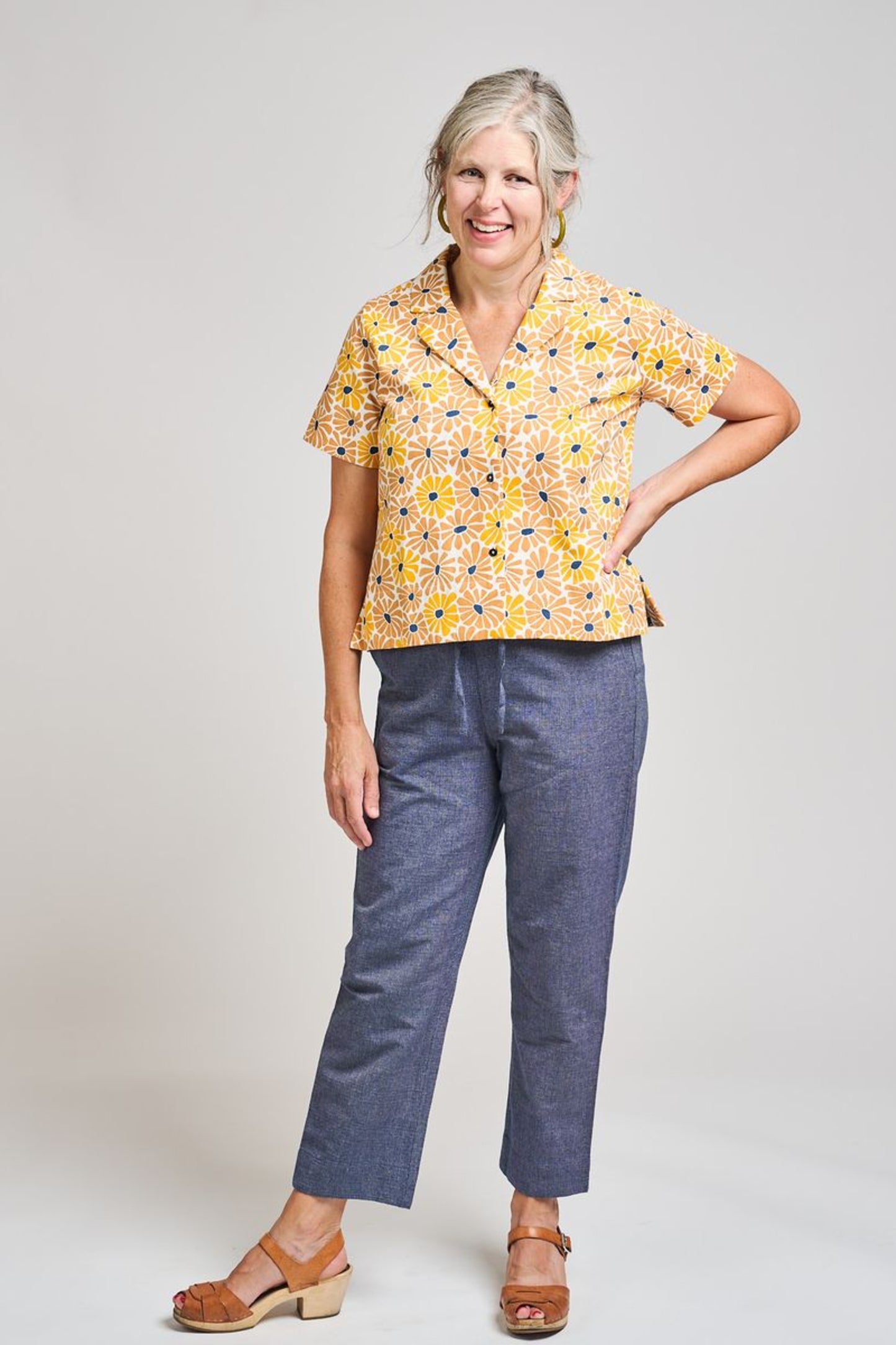 Image of model wearing MATA TRADERS Fair Trade Certified Camp Shirt in Sunny Floral Print. Camp Top is an open neck vintage inspired short sleeve button down. Sunny Floral Print is an orange, yellow, and teal sunflower motif hand block printed on Cotton twill fabric.