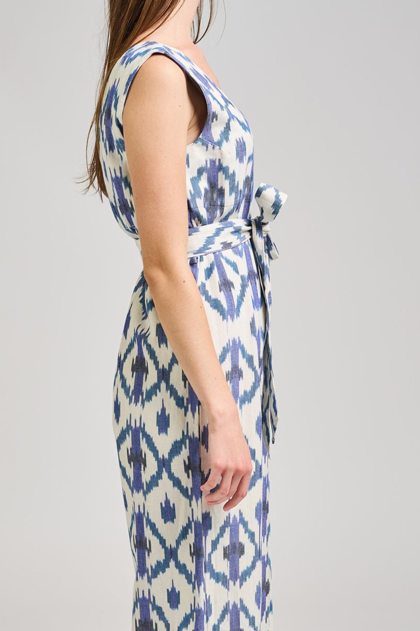 Image of model wearing MATA Traders Fair Trade Certified Jana Jumpsuit in Seashore Ikat. Jana Jumpsuit is a V-Neck and V-Back sleeveless jumpsuit with a straight/wide leg and removable sash belt. Seashore Ikat is a handwoven natural white Cotton Fabric with purple and teal Ikat motif pattern throughout.
