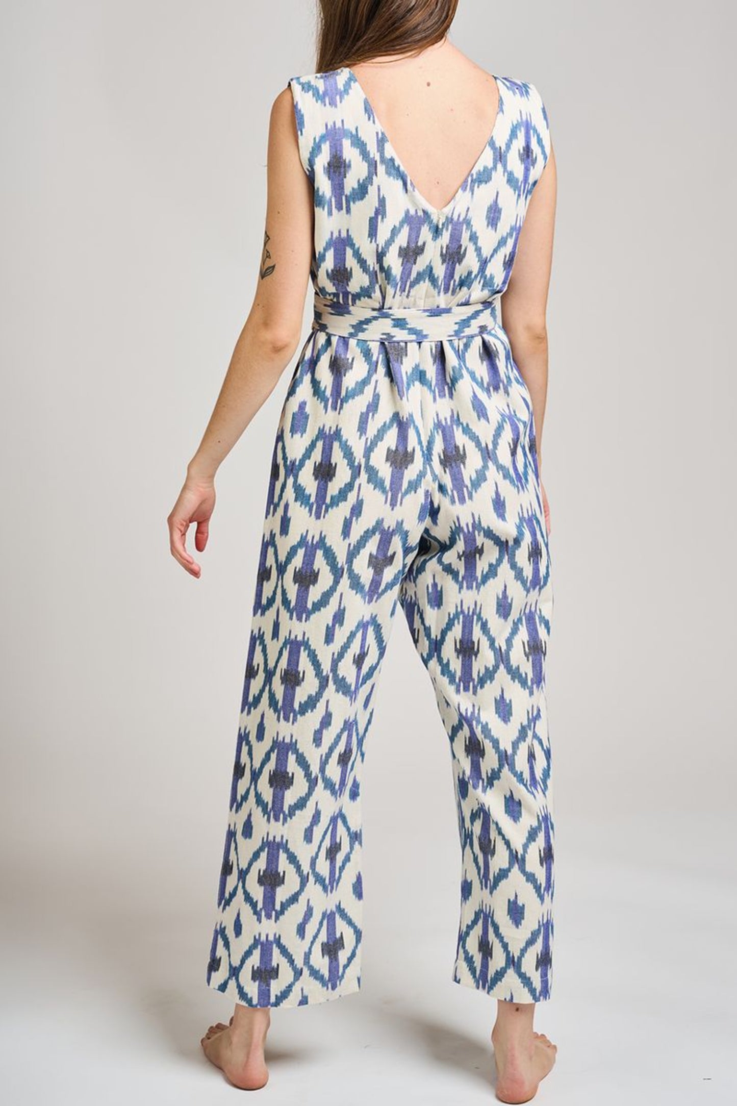 Image of model wearing MATA Traders Fair Trade Certified Jana Jumpsuit in Seashore Ikat. Jana Jumpsuit is a V-Neck and V-Back sleeveless jumpsuit with a straight/wide leg and removable sash belt. Seashore Ikat is a handwoven natural white Cotton Fabric with purple and teal Ikat motif pattern throughout.