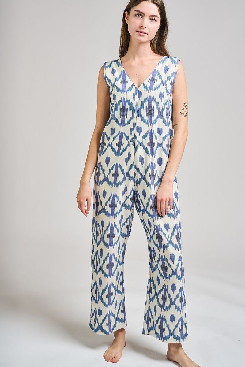 Image of model wearing MATA Traders Fair Trade Certified Jana Jumpsuit in Seashore Ikat. Jana Jumpsuit is a V-Neck and V-Back sleeveless jumpsuit with a straight/wide leg and removable sash belt. Seashore Ikat is a handwoven natural white Cotton Fabric with purple and teal Ikat motif pattern throughout.
