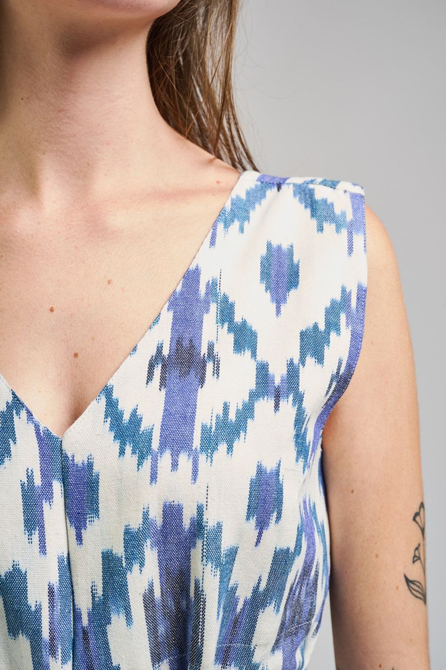 Image of model wearing MATA Traders Fair Trade Certified Jana Jumpsuit in Seashore Ikat. Jana Jumpsuit is a V-Neck and V-Back sleeveless jumpsuit with a straight/wide leg and removable sash belt. Seashore Ikat is a handwoven natural white Cotton Fabric with purple and teal Ikat motif pattern throughout.