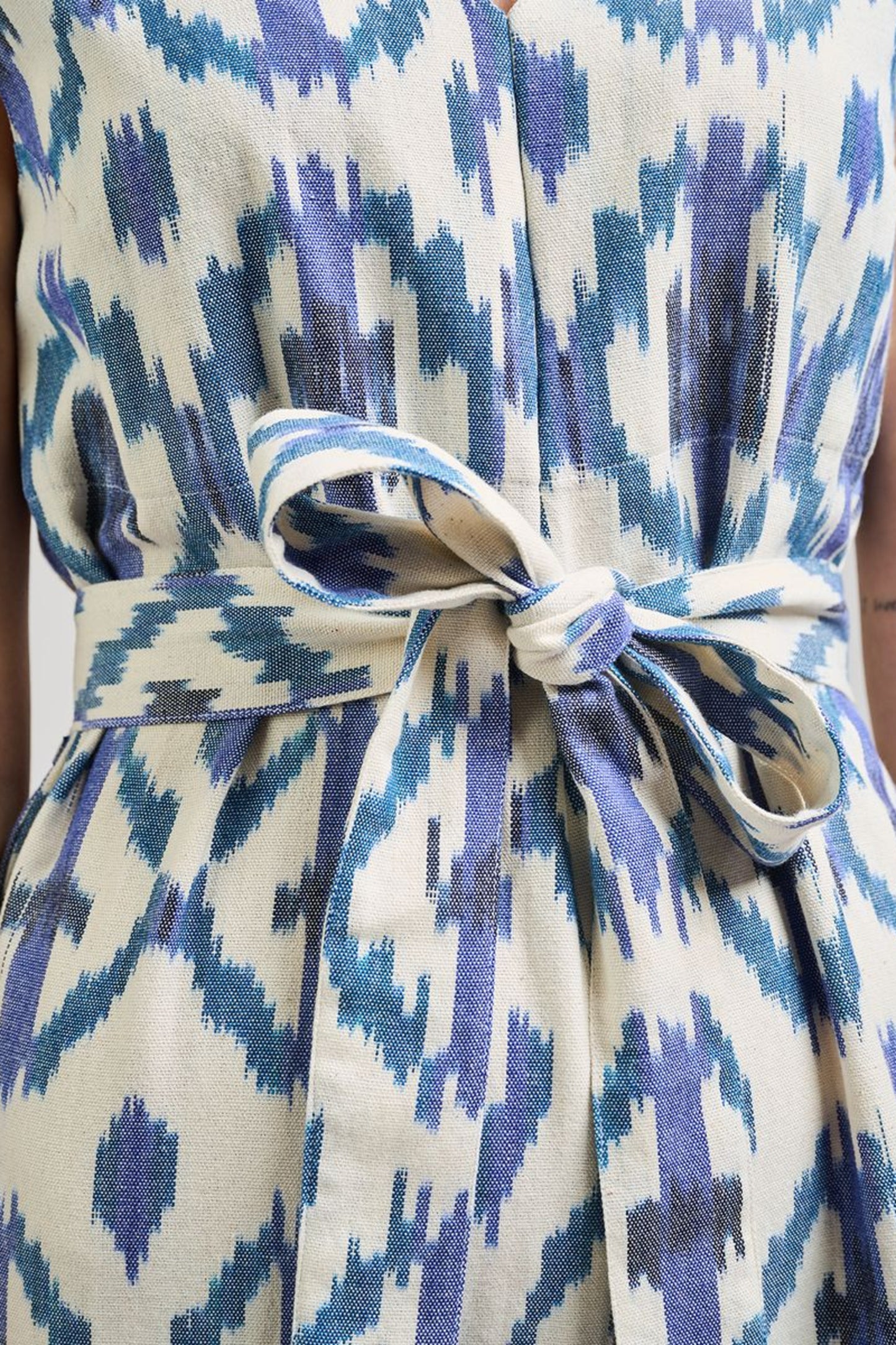 Image of model wearing MATA Traders Fair Trade Certified Jana Jumpsuit in Seashore Ikat. Jana Jumpsuit is a V-Neck and V-Back sleeveless jumpsuit with a straight/wide leg and removable sash belt. Seashore Ikat is a handwoven natural white Cotton Fabric with purple and teal Ikat motif pattern throughout.