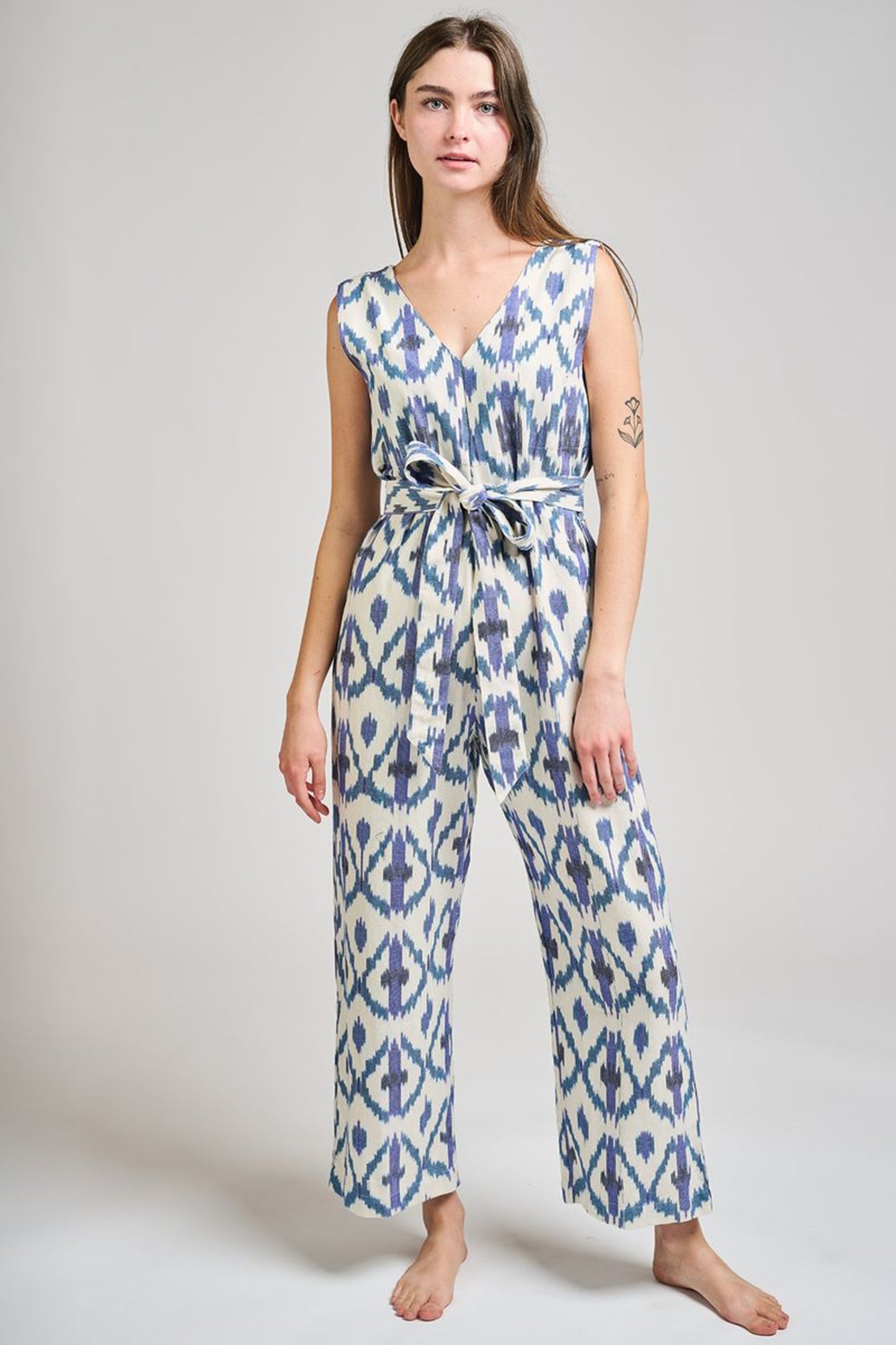 Image of model wearing MATA Traders Fair Trade Certified Jana Jumpsuit in Seashore Ikat. Jana Jumpsuit is a V-Neck and V-Back sleeveless jumpsuit with a straight/wide leg and removable sash belt. Seashore Ikat is a handwoven natural white Cotton Fabric with purple and teal Ikat motif pattern throughout.