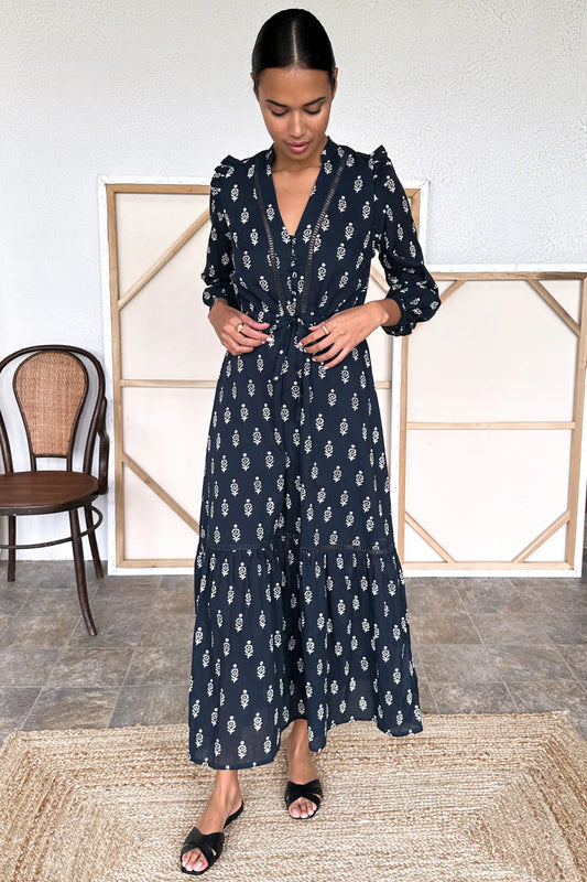 Image of model wearing Emerson Fry Frances V Dress in Margeurite Print. Frances is a Maxi length Bohemian inspired dress with open v-neckline, slight puff shoulder, long sleeve, cinchable tie waist, and Maxi length tiered skirt. Marguerite print is a geometric white Ikat on Navy blue.