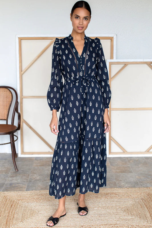 Image of model wearing Emerson Fry Frances V Dress in Margeurite Print. Frances is a Maxi length Bohemian inspired dress with open v-neckline, slight puff shoulder, long sleeve, cinchable tie waist, and Maxi length tiered skirt. Marguerite print is a geometric white Ikat on Navy blue.