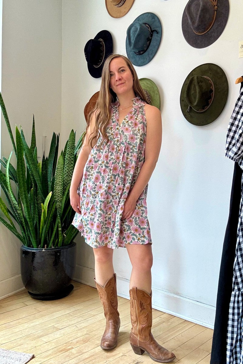 Image of model wearing MAELU DESIGNS Poppy Mini Dress in Cici Print. Poppy is a sleeveless cotton sundress that hits about the knee with an a-line, tiered body and ruffled collar. Offered in a block printed Cotton featuring an all over purple, teal, and yellow floral print. 