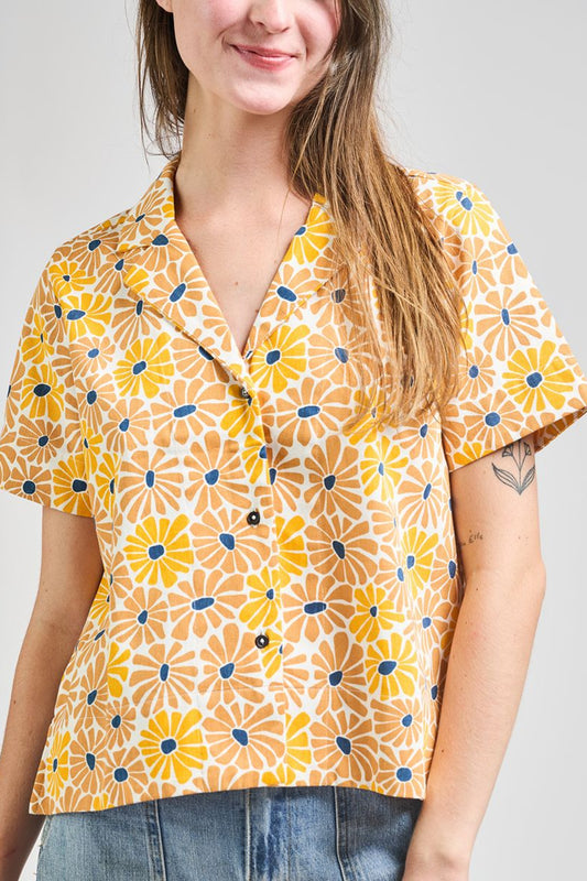 Image of model wearing MATA TRADERS Fair Trade Certified Camp Shirt in Sunny Floral Print. Camp Top is an open neck vintage inspired short sleeve button down. Sunny Floral Print is an orange, yellow, and teal sunflower motif hand block printed on Cotton twill fabric.