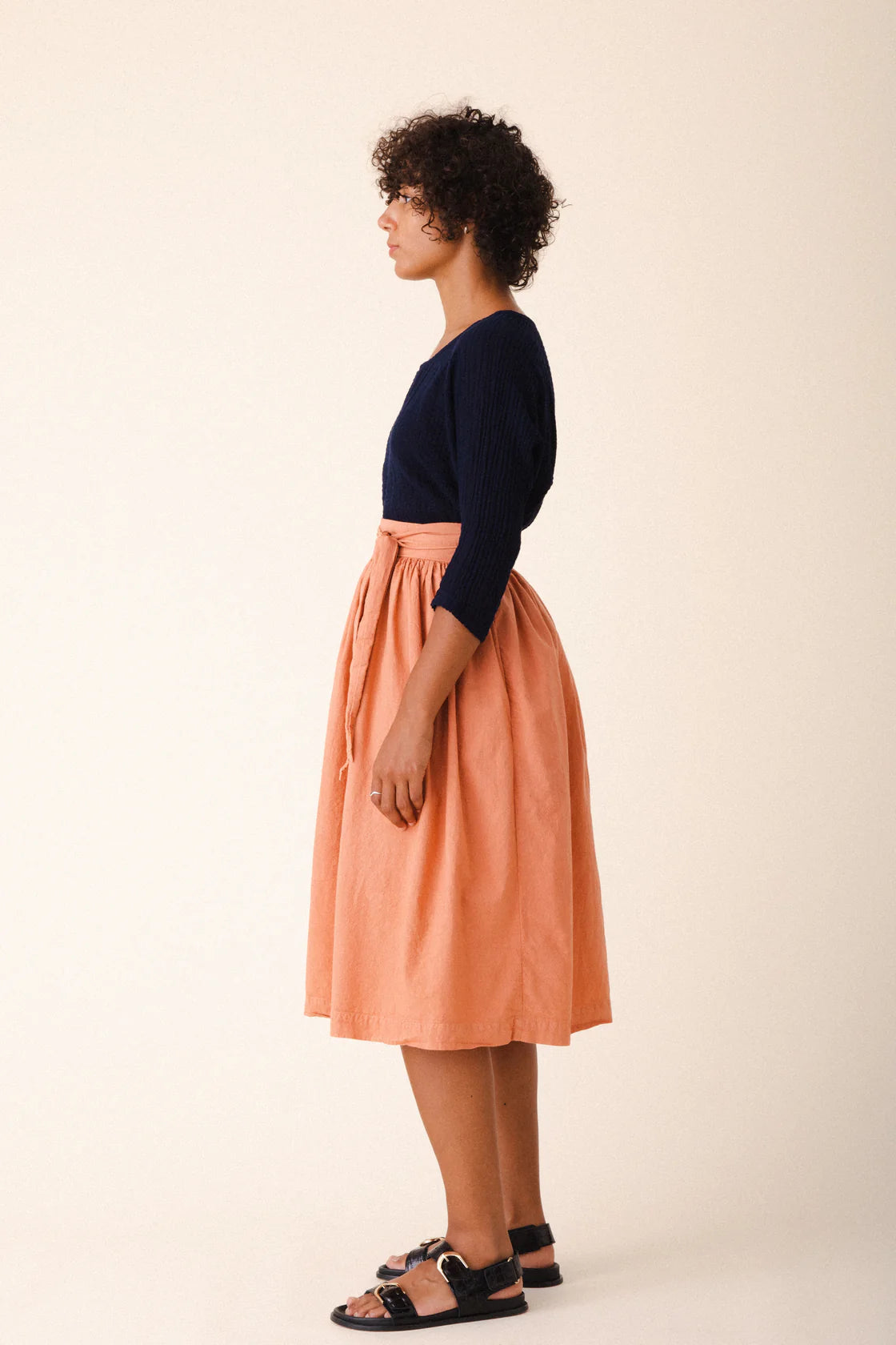 Image of model wearing Prairie Underground Interval Wrap Skirt in Cedar Chest. Interval Skirt is a true wrap skirt that hits below the knee with a sash tie, made in Seattle, Washington from a Cedar Pink organic cotton muslin.