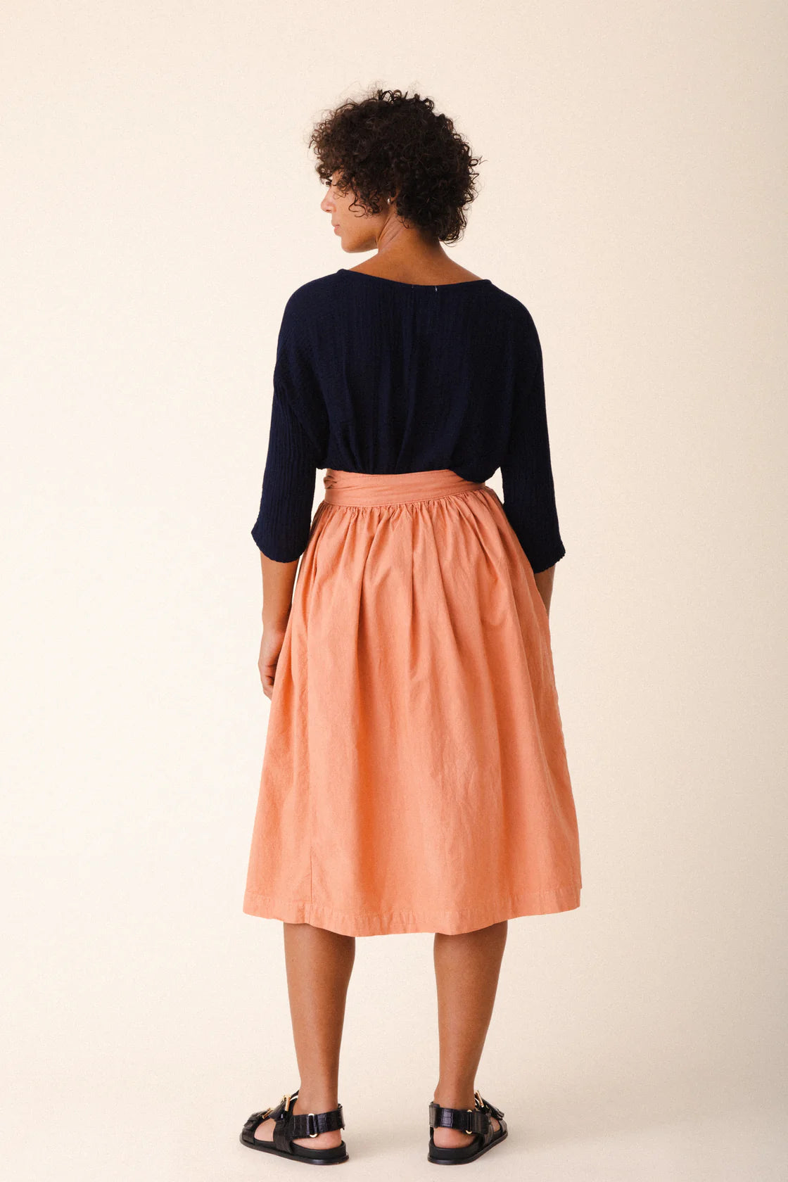 Image of model wearing Prairie Underground Interval Wrap Skirt in Cedar Chest. Interval Skirt is a true wrap skirt that hits below the knee with a sash tie, made in Seattle, Washington from a Cedar Pink organic cotton muslin.